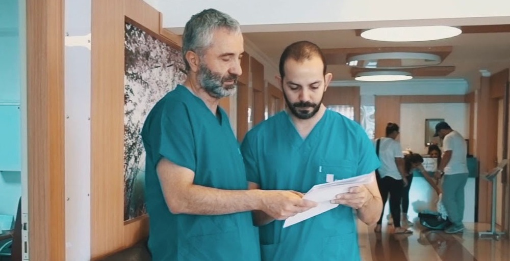 Due medici con camice verde consultano una cartella clinica nel corridoio luminoso del Gynolife IVF Center, con pazienti e personale sullo sfondo.