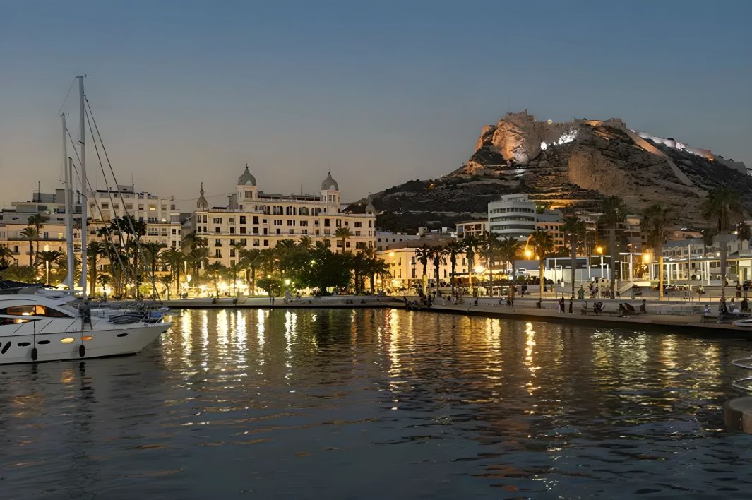 Veduta serale del porto di Alicante con yacht ormeggiati, lungomare illuminato e Castello di Santa Barbara sulla collina sullo sfondo.
